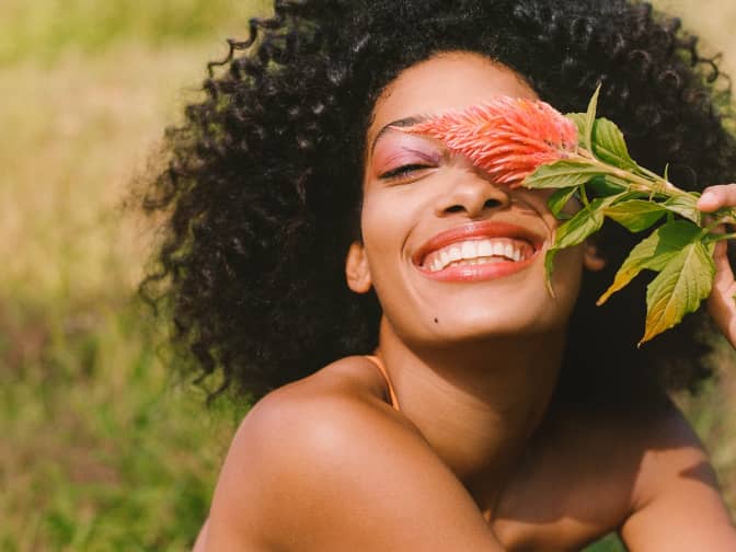 African american woman smiling and delicately holding a single flower over her eye in the sunlight
