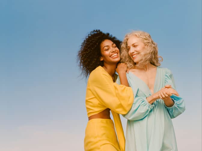 Curly haired Black woman in a yellow dress smiling and resting her hand on the shoulder of a curly haired white woman smiling in a blue dress