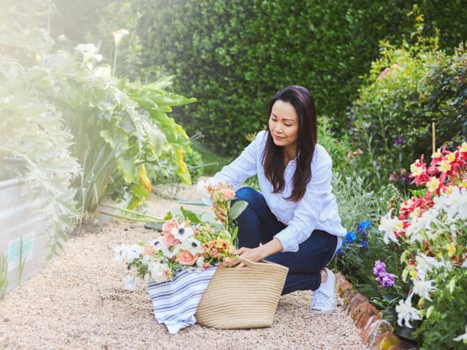 woman kneeling in garden placing flowers into a woven reusable shopping tote