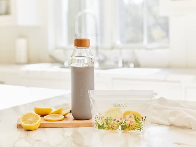 glass water bottle with gray silicone sleeve and wooden cap arranged on the counter top with a sliced lemon and a reusable food bag