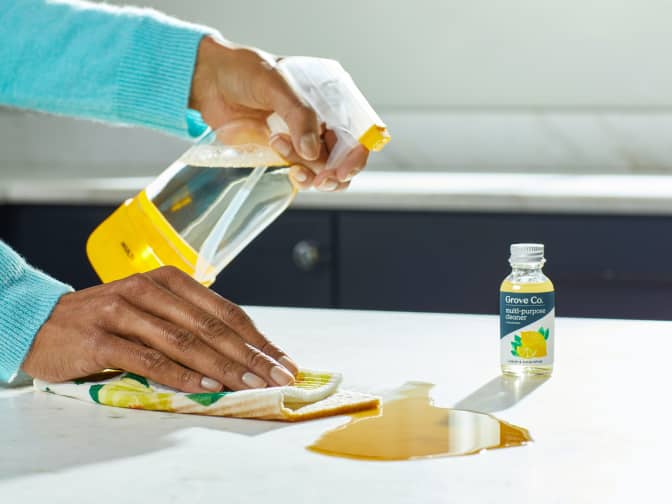 Hand cleaning up a spill with a Grove Co reusable glass spray bottle with yellow slide and snap sleeve, a reusable Grove Co euro dish cloth, and Grove Co multipurpose cleaner concentrate