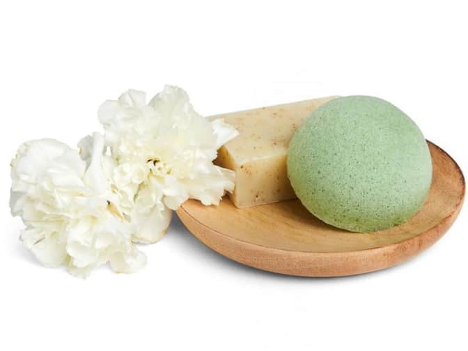 Image of green konjac sponge on wooden plate next to bar soap and white flowers