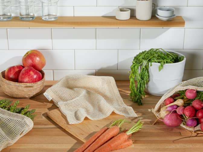 Kitchen counter with reusable produce bags and fresh produce including carrots, radishes, asparagus, and apples.
