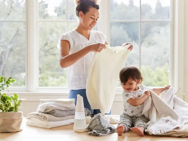 A person folds laundry with natural detergents while a young baby helps