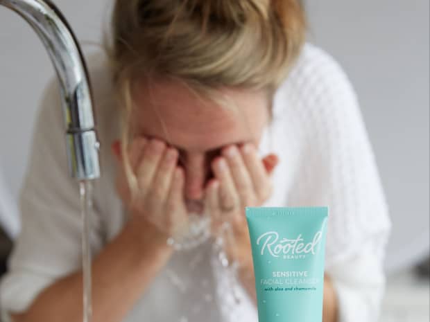 Image of woman washing her face with water at sink with Rooted Beauty skincare product in front of her