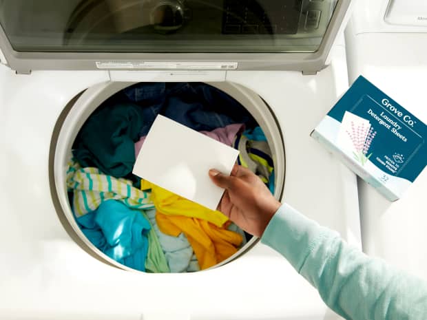 Image of a hand holding a laundry detergent sheet up in front of full load of clothes in washing machine
