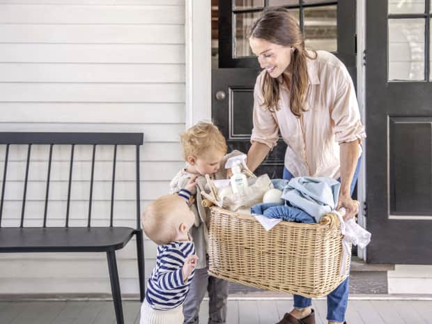 Image of woman holding wicker laundry basket with laundry, stain remover bottle, wool dryer ball with 2 kids peeking inside the basket