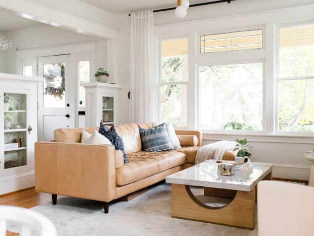 light brown leather couch with throw pillows in a white living room with large windows and a marble coffee table
