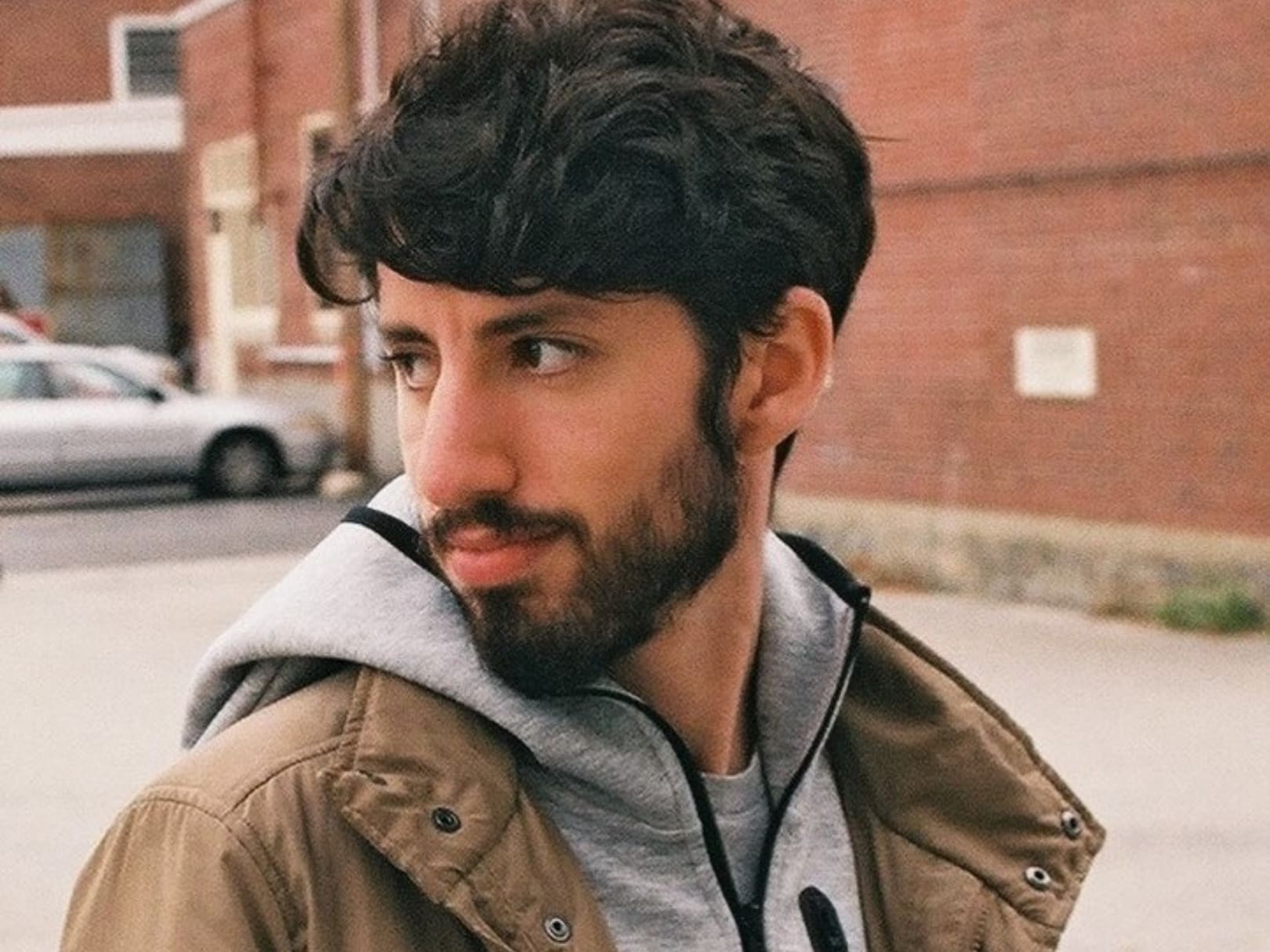 Image of male with dark hair, beard, mustache standing outside in grey hoodie and tan jacket
