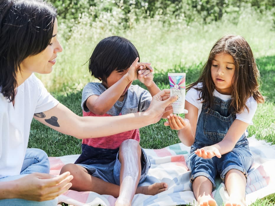 Image of mother putting sunscreen into child's hand while sitting outside on picnic blanket